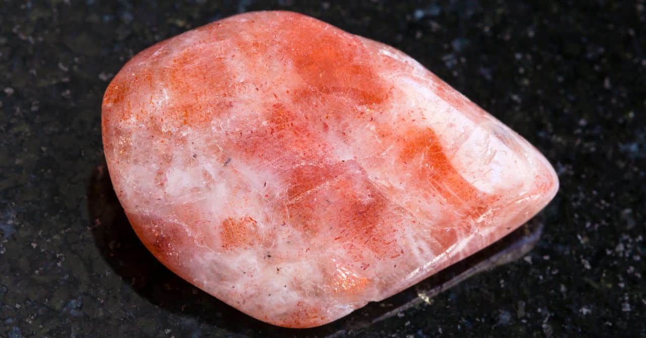 a polished sunstone with its characteristic orange and reddish hues, resting on a dark, reflective surface.