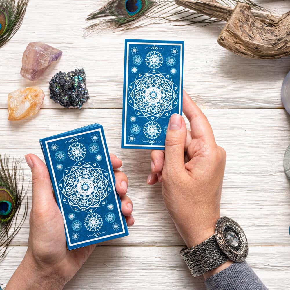 A woman's hands holding a deck of blue tarot cards over a table with crystals and peacock feathers