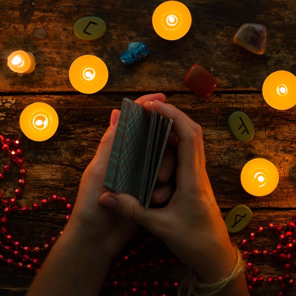 A woman's hands shuffling a deck of turquoise plaid tarot cards over a table with candles, stones, and a bead necklace