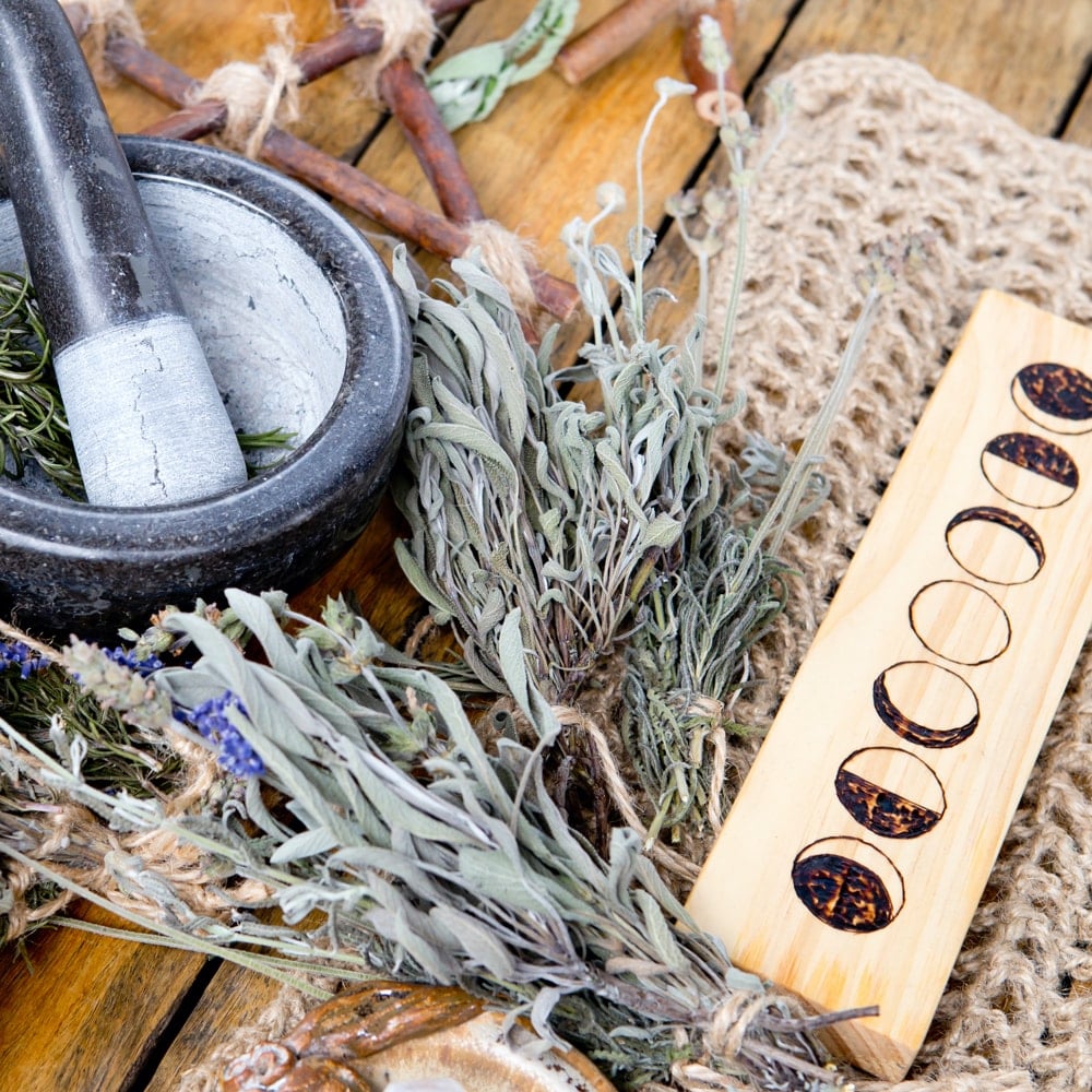 A mortar and pestle on a wooden table with bundles of sage and other herbs