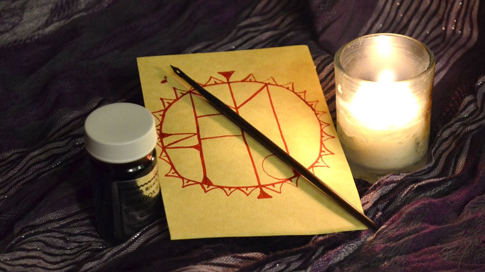 A sigil drawn on paper with a quill and red ink. A lit candle sits beside it