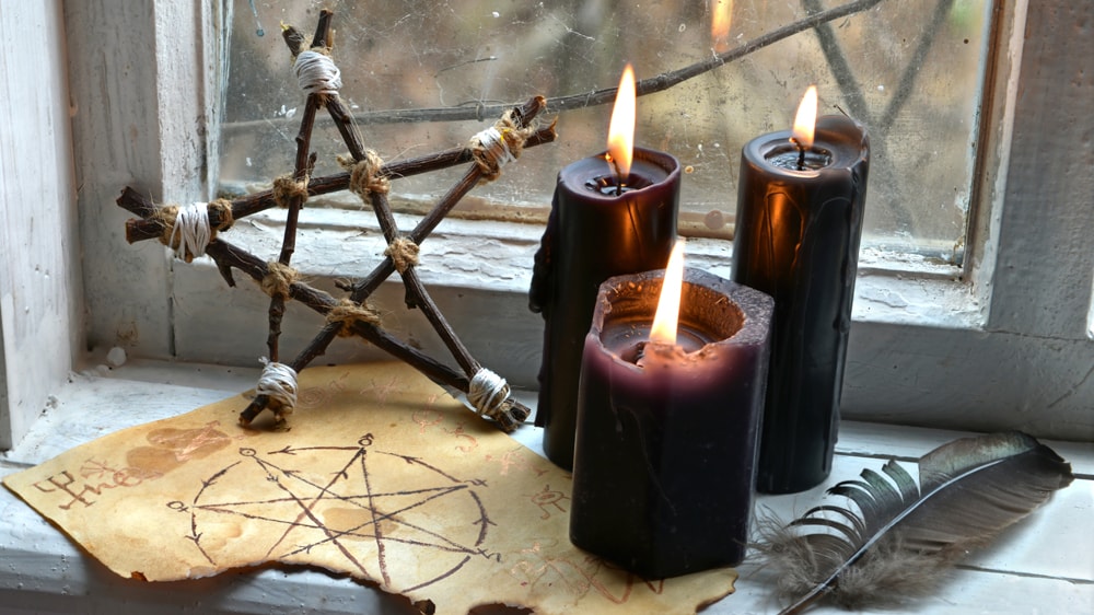 Black candles on an old window sill witha stick pentacle, a feather, and a book of shadows page