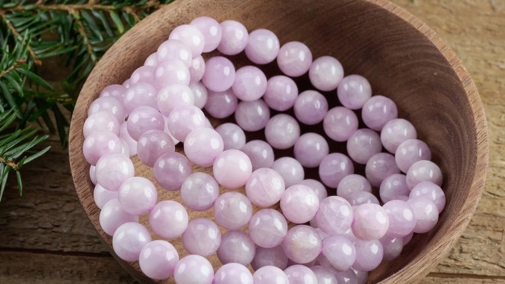 Four rose quartz crystal bead bracelets in a wooden bowl beside some evergreen branches