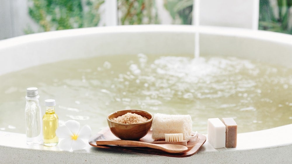 A bath filling while oils, salts, soap, and brushes sit on the edge