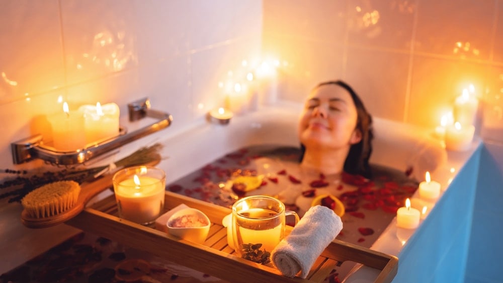 A woman relaxing in a bath filled with rose petals with a glass of tea surrounded by candles