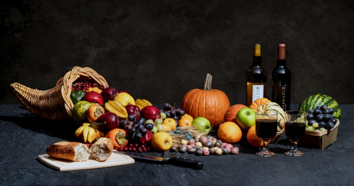 A cornucopia overflowing with harvest fruits and vegetables beside bottles of wine and a loaf of bread