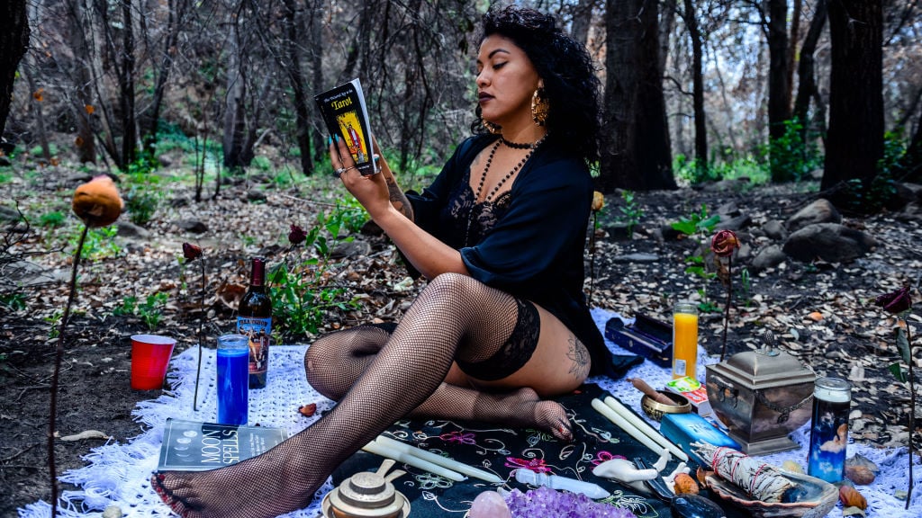 A woman reading a book about tarot in the forest on a blanket with candles, crystals, sage, and other magickal items