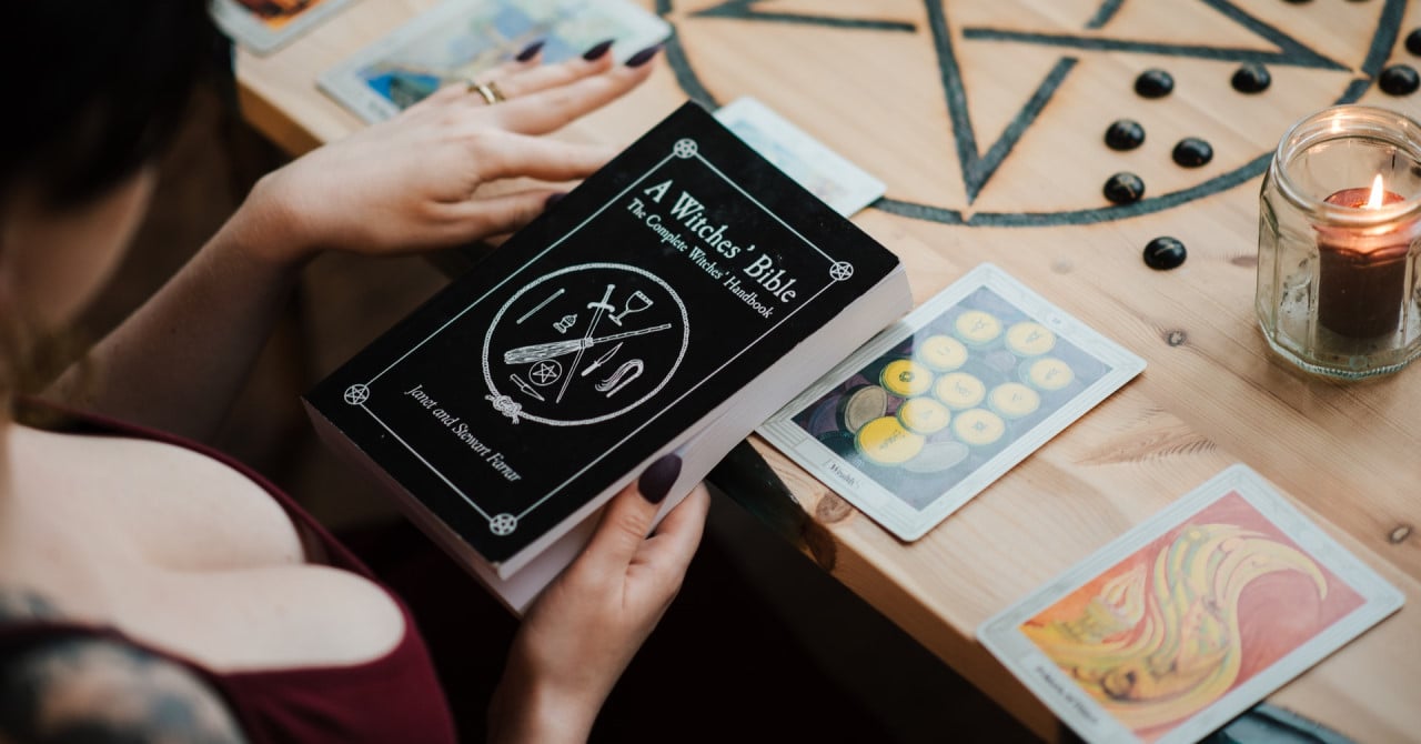 A woman reading a witchcraft book with a candle, tarot cards, and runes on the table in front of her. The table features a pentacle design