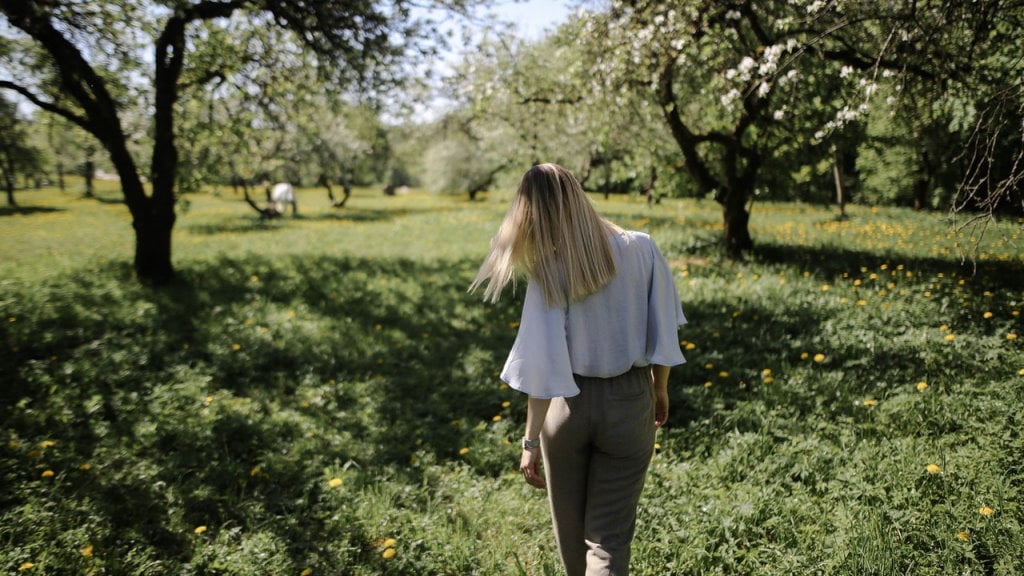 A woman walking through a group of trees in a meadow with flowers starting to bloom