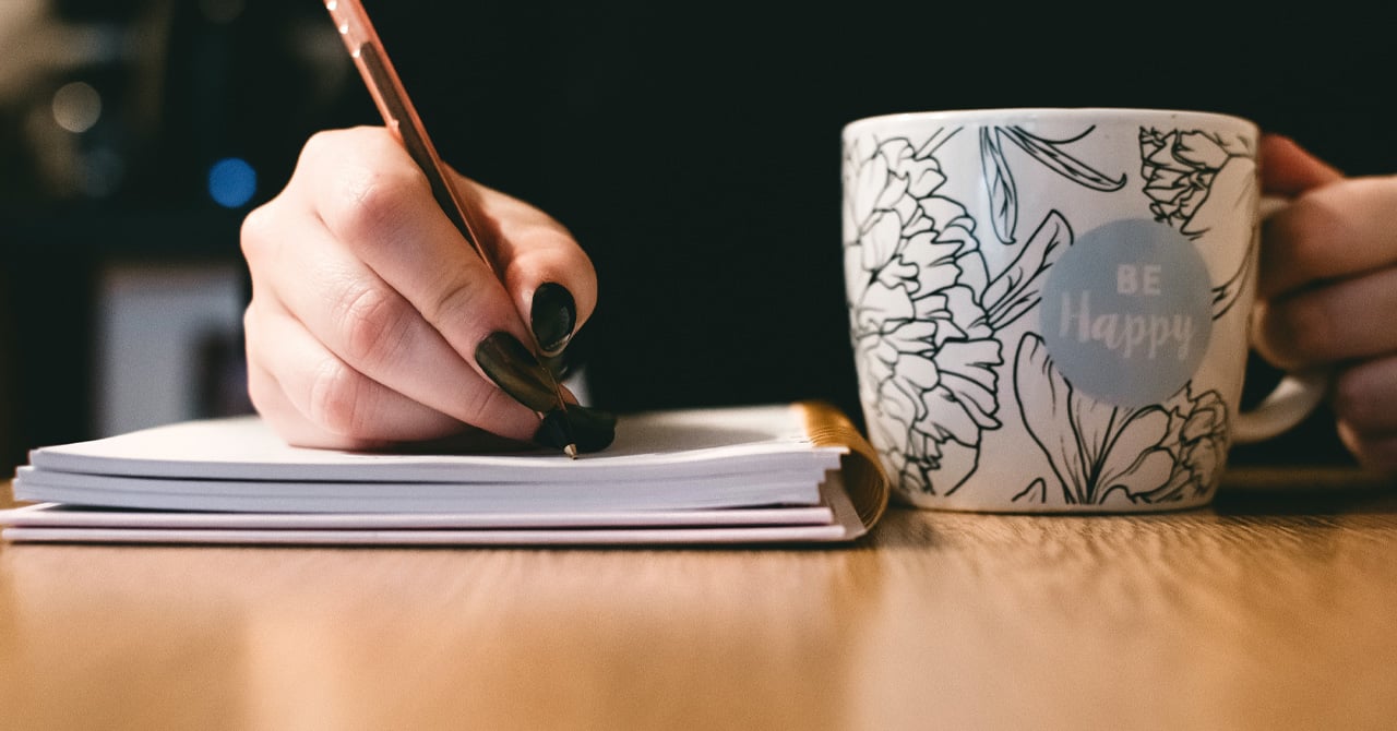 A person writes on a notebook while holding a mug of coffee