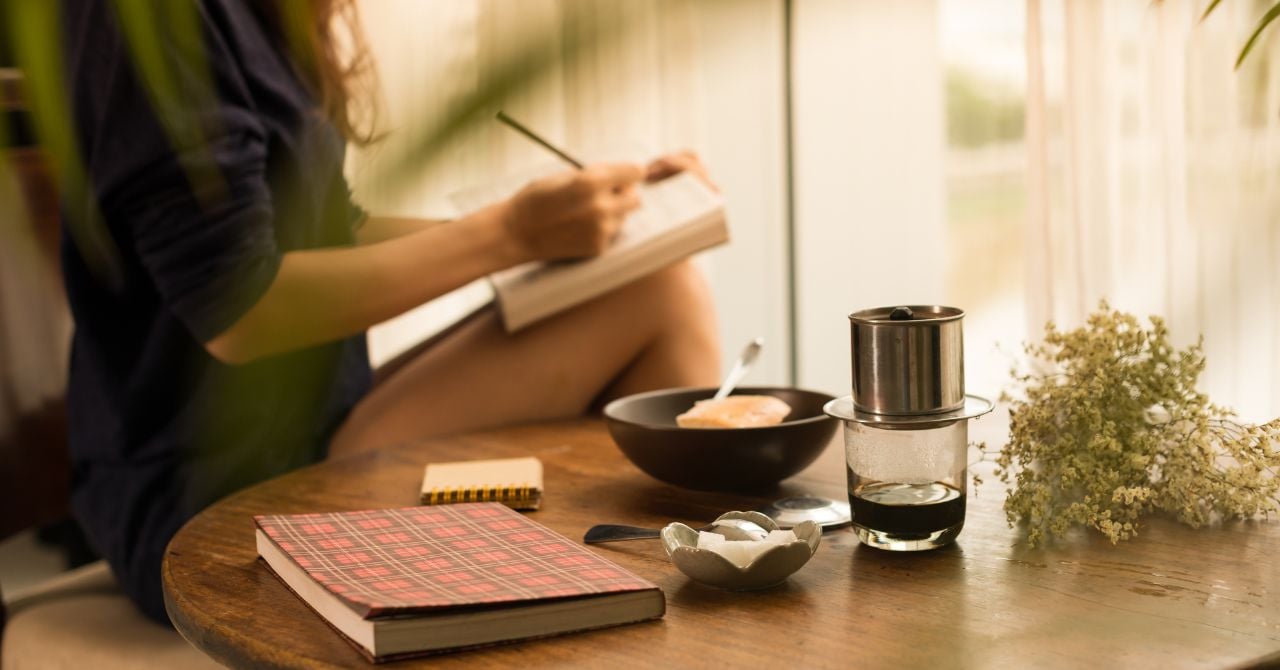 a girl writing on a notebook in front of cup and plate on the table